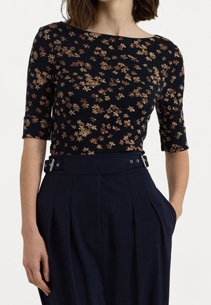 Kvinde iført en sort tætsiddende top med små brune blomsterprint og marineblå højtaljede bukser med spændedetaljer, stående med den ene hånd i lommen.