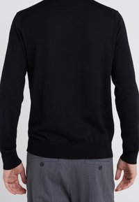 Svart stickad tröja med långa ärmar, ribbade muddar och nederkant, i kombination med grå byxor med knappdetaljer på fickorna.