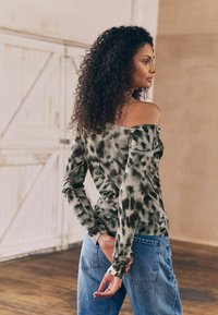 Off-shoulder top i grøn tie-dye mønster med lange ærmer, parret med lyseblå højtaljede jeans. Blødt, strækbart stof.