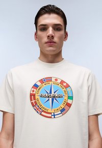 Camiseta de cuello redondo color blanco roto con un diseño de compás colorido que incluye banderas y el texto "CONÉCTATE CON TU VERDADERO YO" y "NAPAPIJRI."