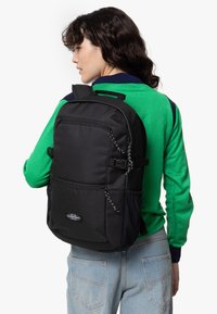 Mochila negra de nailon con tirantes ajustables, bolsillo con cremallera en la parte frontal y acentos decorativos de cordón; llevada en la espalda por una persona que viste un suéter verde.