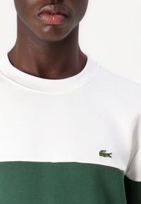 Sudadera blanca y verde con un cuello redondo, que presenta un diseño de bloques de color horizontal y un pequeño logotipo de cocodrilo bordado.