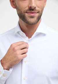 Witte button-up shirt met een spread collar; gladde textuur; zichtbare knoopdetails; persoon die de kraag aanpast met een neutrale handbeweging.