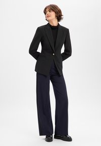 Svart skreddersydd blazer med en enkelt gullknapp, kombinert med vide marineblå bukser. Jevnt stoff, strukturert design og minimale detaljer.