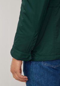 Grön, vattentät jacka med en synlig vikbar mudd och sidogångjärn. Designen är elegant, med en slät textur och minimal detaljer.