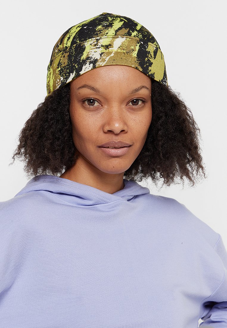 Wielokolorowa opaska na głowę z abstrakcyjnym wzorem w czerni, zieleni i żółci. Model nosi jasnofioletową bluzę z kapturem.