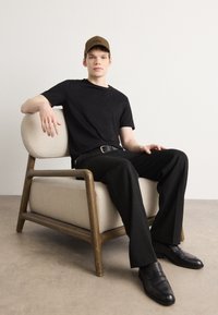 Black t-shirt, black trousers, and brown cap worn by a seated individual on a light beige upholstered chair with wooden legs. Black shoes.