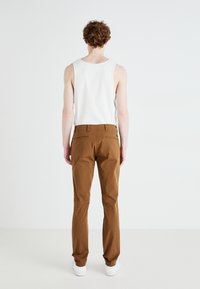 Brune slim-fit bukser med en glat tekstur, baglommer og et logo mærke ved livet, parret med en hvid ærmeløs skjorte.