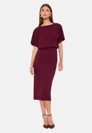 Nő, aki testhez simuló, térdig érő, sötét bordó ruhát visel, laza rövid ujjakkal, arany karika fülbevalókkal, vastag karkötővel és barna, bokapántos magassarkú cipőkkel.