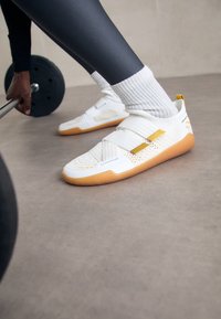 Αθλητής που φορά λευκά αθλητικά παπούτσια με κίτρινες λεπτομέρειες και ριγέ κάλτσες, κρατώντας μια μπάρα με μαύρα βάρη σε μια γυμναστική επιφάνεια.