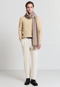 Beiger gerippter Pullover, hellbeige Schal, cremefarbene Hose und schwarze Stiefeletten. Glatte Stoffe mit Fransen Details am Schal.