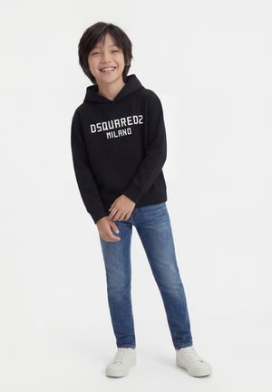 Bambino sorridente con felpa nera DSQUARED2 Milano, jeans blu e sneakers bianche, in piedi davanti a uno sfondo bianco uniforme.