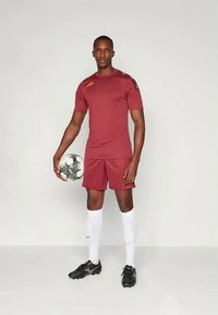 Bordo futbola komplekts ar īsām piedurknēm un šortiem, ar zelta akcentiem. Spēlētājs tur ierobežotu melnbaltu futbola bumbu, vilkdams melnas spārda.