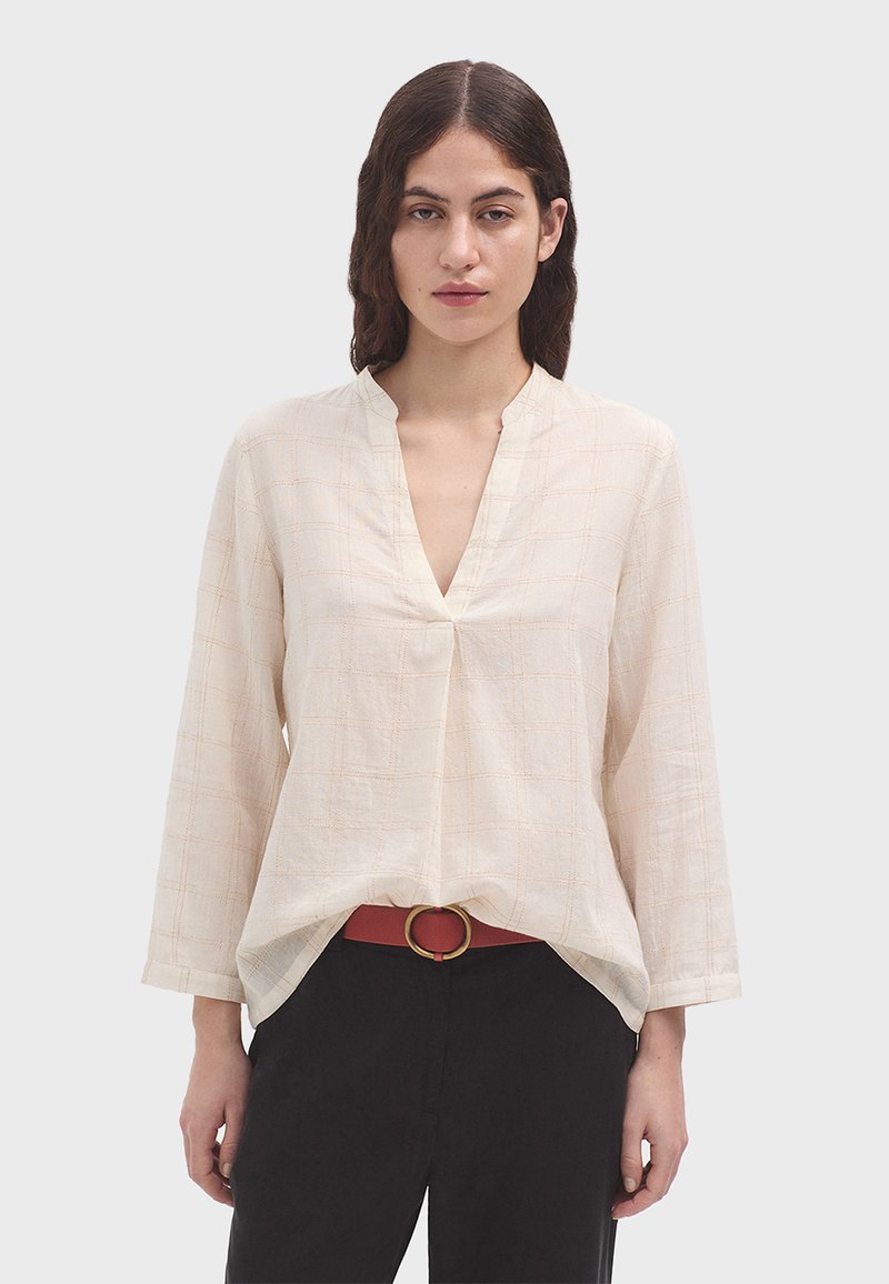 Helle beige Bluse mit langen Ärmeln und V-Ausschnitt, die ein dezentes Rautenmuster aufweist und locker geschnitten ist. Kombiniert mit einem roten Gürtel und dunklen Hosen.