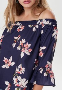 Laivastonsininen olkaimeton toppi kevyestä kankaasta, jossa on vaaleanpunaisia ja valkoisia kukkakuoseja, joustava kaula-aukko ja kellotetut hihansuut.