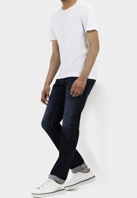 Hombre vistiendo una camiseta blanca lisa, jeans oscuros arremangados, calcetines grises y zapatillas blancas, de pie con las piernas cruzadas contra un fondo blanco.