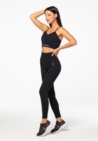 Black sports bra with gathered front, paired with high-waisted black leggings featuring ribbed texture and logo accents. Black sneakers with pink details.