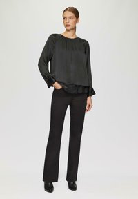 Zwarte satijnen blouse met lange mouwen, een gathered halslijn en kantafwerking, gecombineerd met zwarte uitlopende broek en enkelboots, tegen een lichte achtergrond.