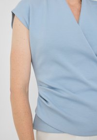 Světle modrý top s krátkým rukávem a překříženým designem, vyrobený z jemné tkaniny. Zřetelný detail na bokách s nařasením a zaoblený lem.