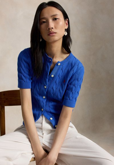 Mlada ženska z dolgimi črnimi lasmi, oblečena v modro kratkoročnu pleteno jopico in bele hlače, sedi na lesenem stolu.