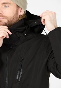Giacca impermeabile nera con colletto alto, tasche con zip e cappuccio regolabile. Texture liscia e dettagli hardware prominenti.