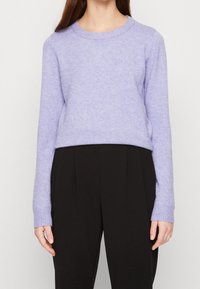 Mujer vistiendo un suéter de punto lila claro y pantalones negros de cintura alta, de pie frente a un fondo claro y sencillo.