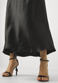 Svart, knelengde skjørt laget av teksturert stoff, parret med svarte sandaler med stiletthæl og ankelremmer.