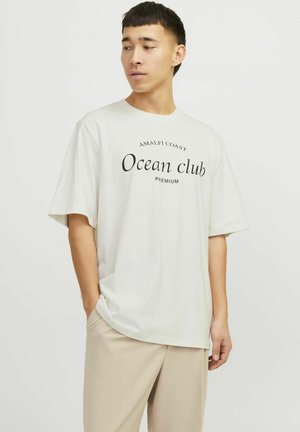 Svijetlo bež pamučna majica s kratkim rukavima, s crnim natpisom "AMALFI COAST Ocean club PREMIUM" u zakrivljenom dizajnu.