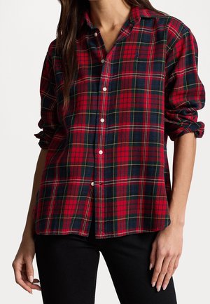 Vrouw die een geruit overhemd in rood, marineblauw en geel draagt met opgerolde mouwen en een zwarte broek, staand tegen een effen achtergrond.