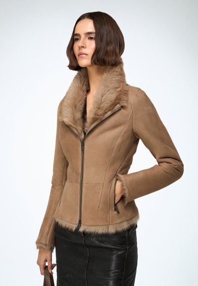 Veste en daim marron avec col et poignets en fausse fourrure, coupe ajustée, fermeture éclair et poches latérales. Portée par-dessus une jupe noire texturée.