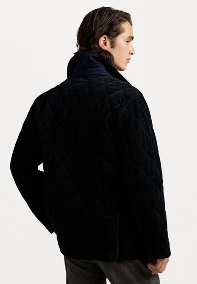 Melna kvilota jaka no kordūru auduma, ar augstu apkakli, ribota tekstūra un sānu kabatām, attēlota no sānu leņķa.