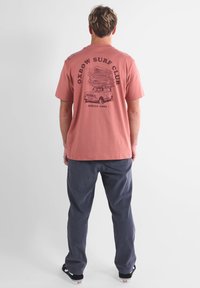 T-shirt rose en coton à manches courtes avec un motif de camion et de planches de surf au dos. Associé à un pantalon bleu foncé et des chaussures noires.