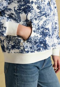 Ręka w kieszeni białego hoodie z niebieskim nadrukiem zwierząt i drzew, w połączeniu z niebieskimi dżinsami na neutralnym tle.