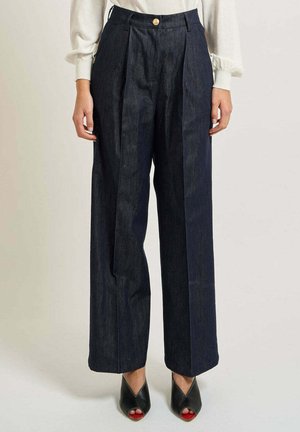 Donna che indossa pantaloni di denim scuro a vita alta e gamba larga con un bottone dorato e scarpe nere aperte con tacco.