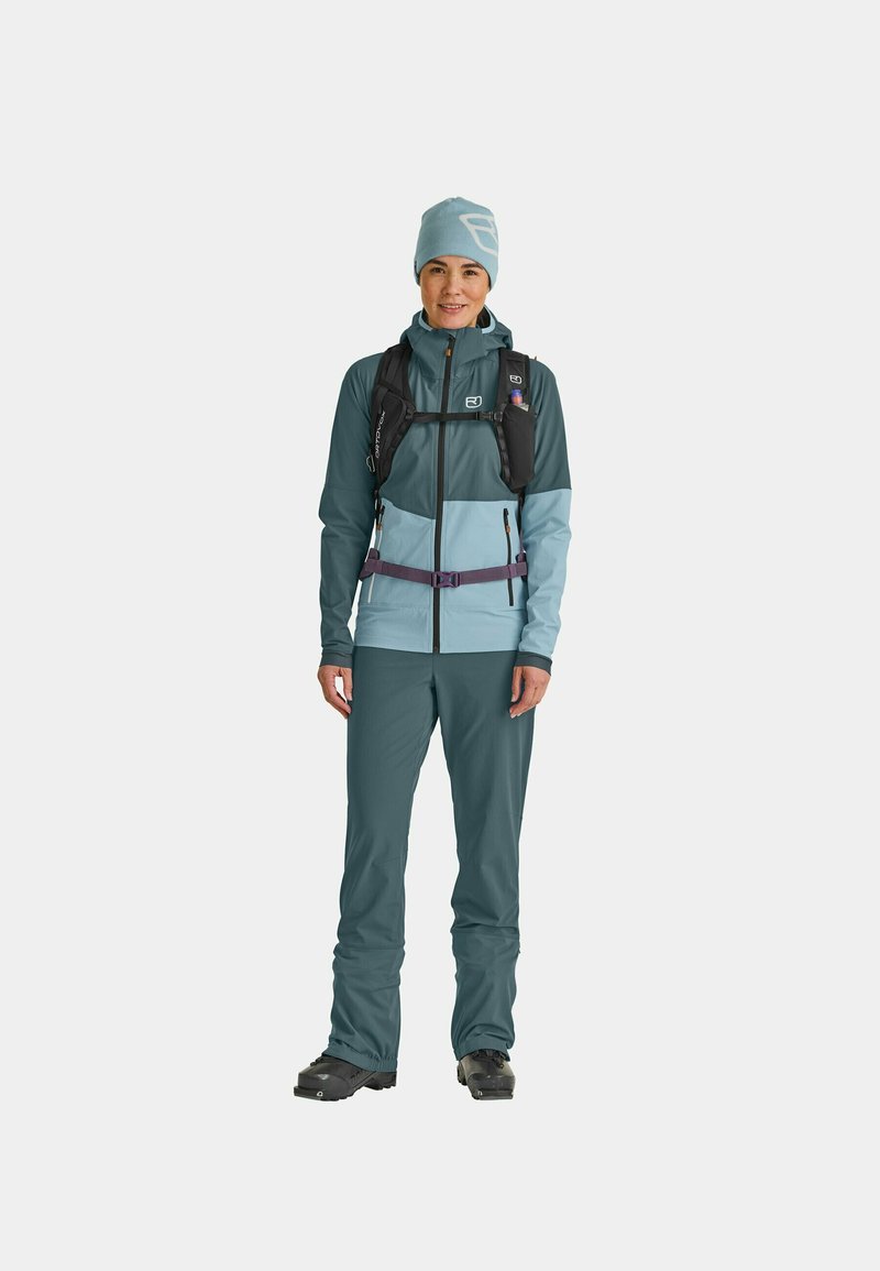 Teal and light blue outdoor outfit, featuring a zip-up jacket with hood and matching pants. Includes a black hydration pack and beanie.
