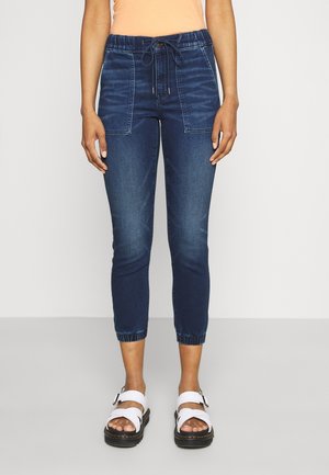 Femme portant un pantalon jogger en denim bleu foncé court avec poches avant, poignets élastiques et sandales blanches à boucle, debout devant un fond uni.