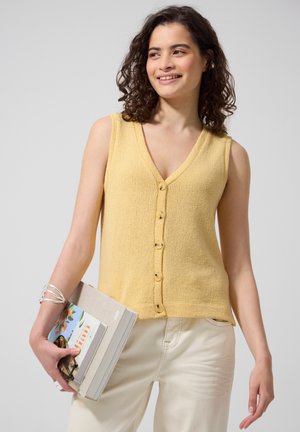 Femme aux cheveux bouclés portant un haut sans manches jaune boutonné et un pantalon crème, tenant des livres et souriant en regardant sur le côté.