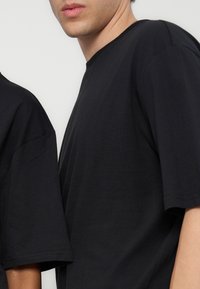 Két ember részleges látképe, akik egyszínű fekete rövid ujjú pólót viselnek világos háttér előtt.