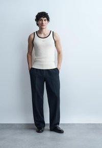 Ung mann med krøllete hår iført hvit singlet, svarte vide bukser og svarte sko, stående med hendene i lomma mot ensfarget bakgrunn.