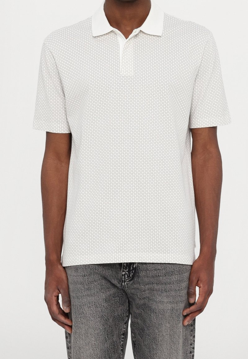 Homme portant un polo blanc à manches courtes avec un petit motif circulaire et un jean gris délavé, debout devant un fond uni.