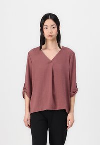 Løs-fittende V-hals mauve bluse med tre kvartlange ermer, som har rullede mansjetter og et mykt, teksturert stoff. Kombinert med svarte bukser.