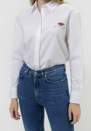 Kobieta ubrana w białą, długorękawową koszulę na guziki z małym czerwono-czarnym logo na piersi, zestawioną z niebieskimi dżinsami z wysokim stanem.