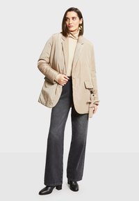 Béžový polstrovaný blazer s klopami, bočním kapsou a detailem na knoflíky. Nositý přes krémový rolák a kombinovaný s tmavými širokými džínami.