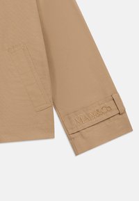 Μανίκι μπεζ σακακιού με ραμμένη τσέπη και λουράκι μανικιού κεντημένο με το λογότυπο "MAX&Co." σε λευκό φόντο.