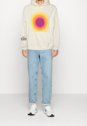 Cream hoodie with a gradient purple and yellow sun design, front pocket, and "studio" text on the sleeve. Paired with light blue jeans and sneakers.