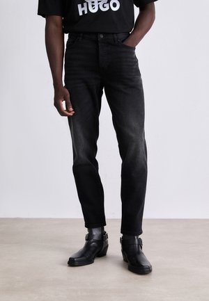 Man wearing black faded jeans, black leather ankle boots with buckles, and a black T-shirt with white "HUGO" logo.
