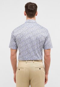 Camicia a maniche corte con un motivo floreale blu e bianco, realizzata in un tessuto leggero, abbinata a pantaloni beige su misura con tasche posteriori.