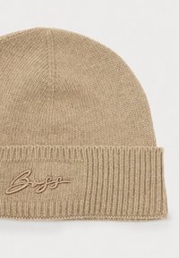 Gorro de punto beige con un puño acanalado y texto bordado. Textura suave, corona redondeada, adecuado para uso casual o clima frío.
