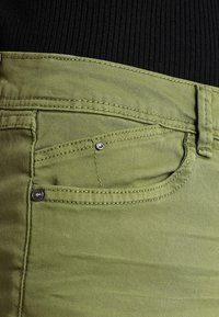Tejido de mezclilla verde con una textura suave, que cuenta con bolsillos frontales, costuras en contraste y acentos de herrajes metálicos.