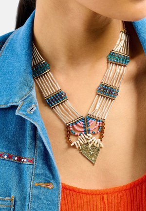 Collier de perles composé de perles en verre multicolores, accents métalliques et un pendentif en forme de cœur orné, présenté sur un jean et un haut orange.